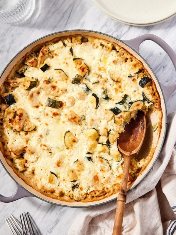 Marble surface with large skillet with cooked chicken zucchini rice casserole and wooden spoon scooping out a serving.