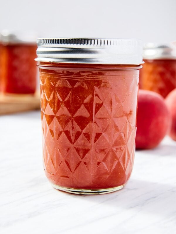 Jam jar filled with orange-colored peach butter sitting on white marble surface with more jam jars in the background.