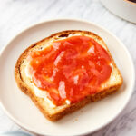 White marble surface with piece of bread spread with butter and peach butter all on white plate with peaches all around.