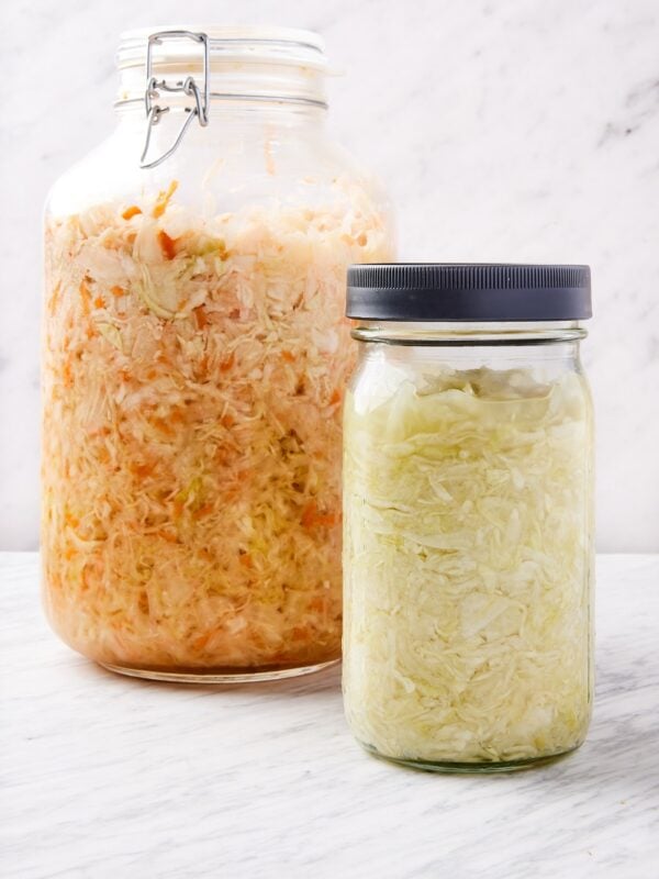 Two jars of homemade sauerkraut sitting on marble surface with white background with one jar filled with carrot along with sauerkraut and the other smaller jar plain.