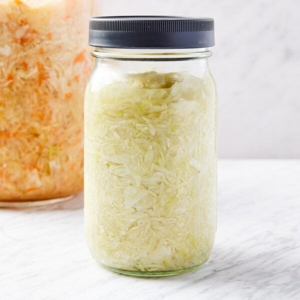 Quart jar filled with homemade sauerkraut made from cabbage with gray screwtop lid with another larger glass jar in background filled with more sauerkraut.