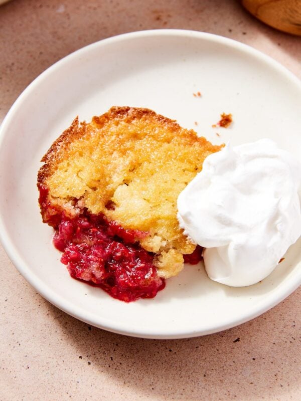 White plate with serving of lazy raspberry cobbler with dollop whipped cream with rest of cobbler and extract raspberries in the background.