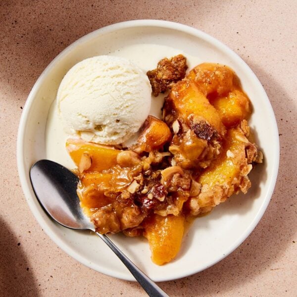 Light pink surface with white plate with serving of peach crisp and scoop of ice cream and spoon resting on plate right before being eaten.