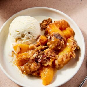 White plate filled with serving of peach crisp with scoop of ice cream melting beside the crisp all on light pink surface.