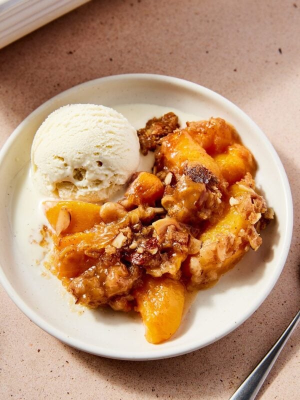 White plate filled with serving of peach crisp with scoop of ice cream melting beside the crisp all on light pink surface.