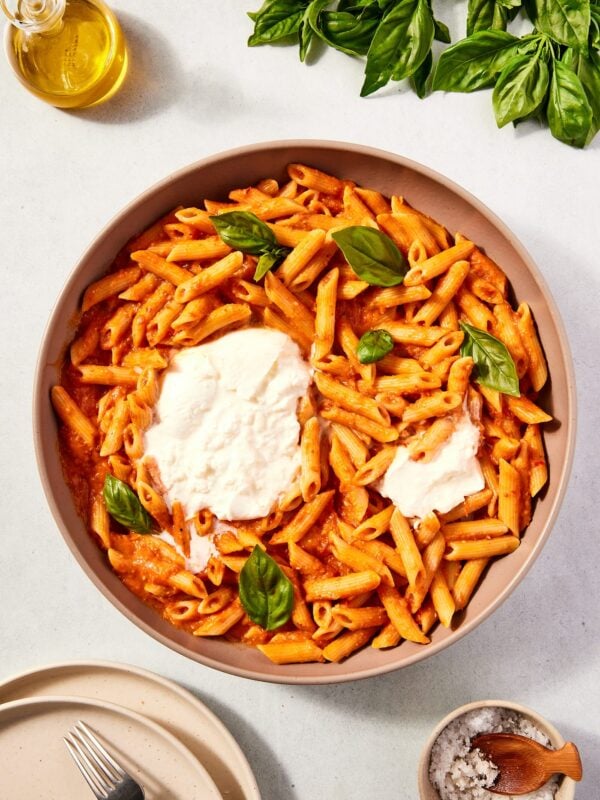 Large bowl serving bowl filled with roasted tomato pasta with large ball of burrata in the middle along with torn basil leaves all on stone surface.