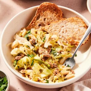 White bowl filled with skillet sausage and cabbage mixture with slices of toasted bread in bowl as well with fork picking up a serving.