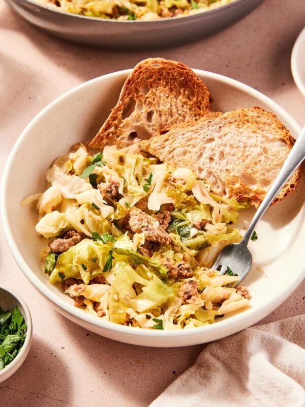 White bowl filled with skillet sausage and cabbage mixture with slices of toasted bread in bowl as well with fork picking up a serving.