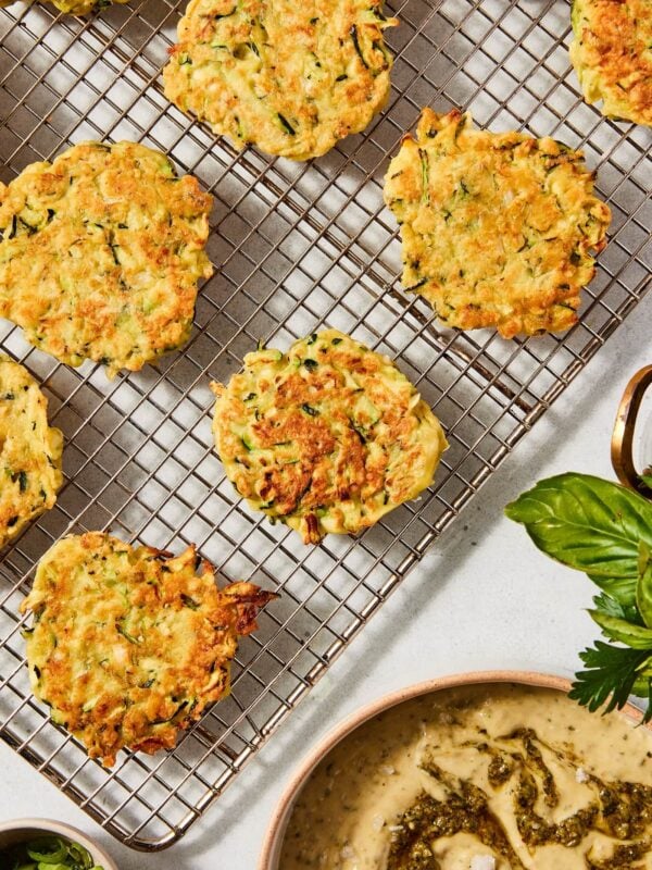 Stone surface with cooling zucchini fritters sitting on wire rack with pesto aioli in stoneware bowl to the side.