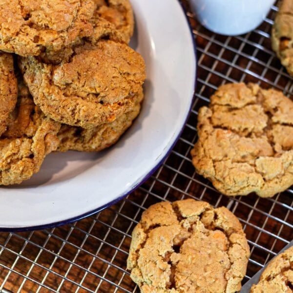 A plate full of cookies sits on a wooden table next to cookies on a wire rack.