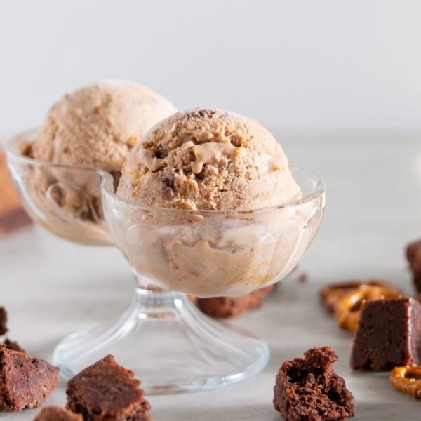 Two scoops of chocolate ice cream sit in a glass container with pretzels and brownies scattered beneath it.