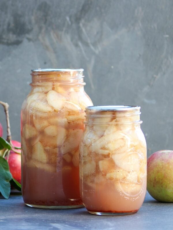 Two glass jars full of apple slices sit next to whole apples.