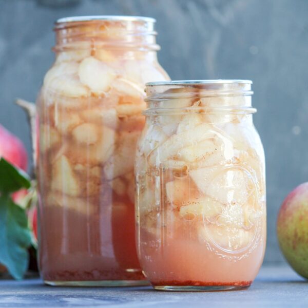 Two glass jars full of apple slices sit next to whole apples.