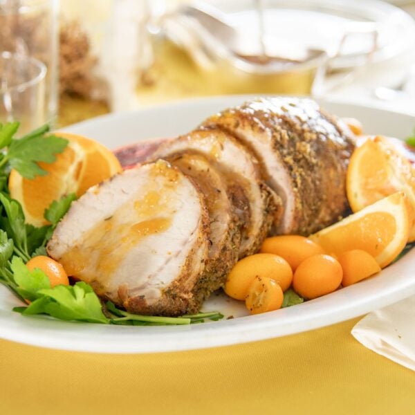 Sliced pork roast garnished with herbs, tomatoes, and oranges sits on a plate.