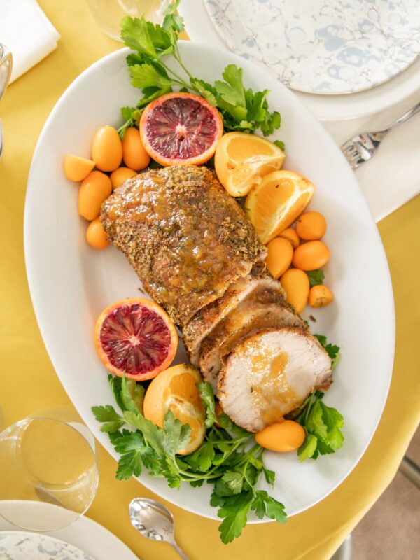 Sliced pork roast sits on a plate adorned with herbs, citrus fruits and tomatoes.