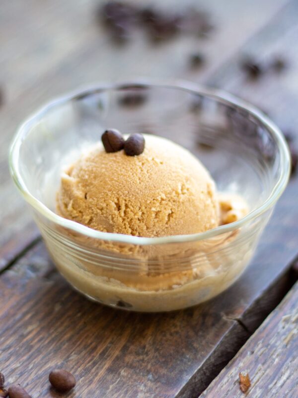 A scoop of ice cream topped with coffee beans rests in a glass dish on a table.