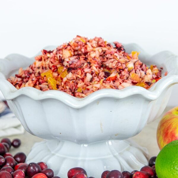Cranberry relish rests in a white decorative dish surrounded by cranberries and fruits.