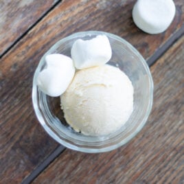 A scoop of ice cream sits next to marshmallows in a glass dish.