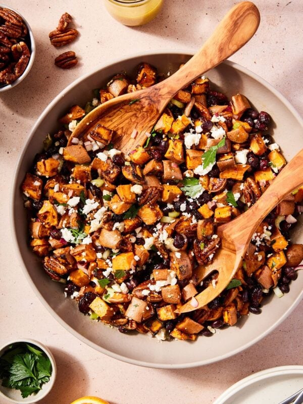 Brown bowl with two wooden tongs scooping up roasted sweet potato salad topped with herbs and feta with more dressing to the side.