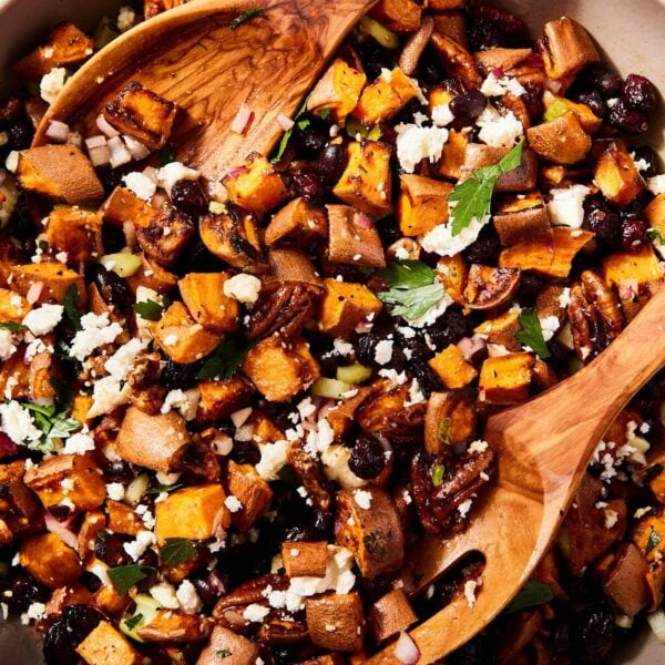Large brown bowl filled with sweet potato salad topped with feta cheese and fresh herbs along with dried cranberries all with wooden tongs about to mix everything together.