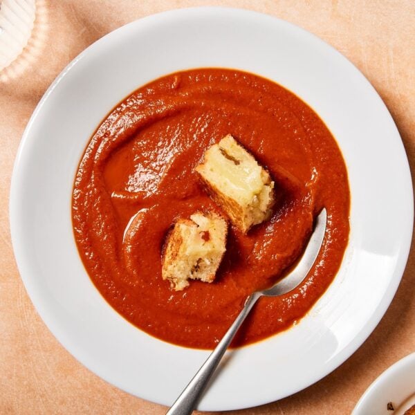 White bowl filled with roasted tomato soup with grilled cheese croutons sitting on surface with spoon scooping some soup.