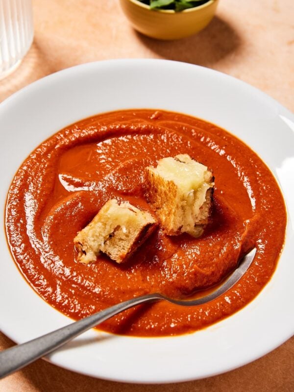 White bowl filled with creamy red colored roasted tomato soup with grilled cheese croutons on top.