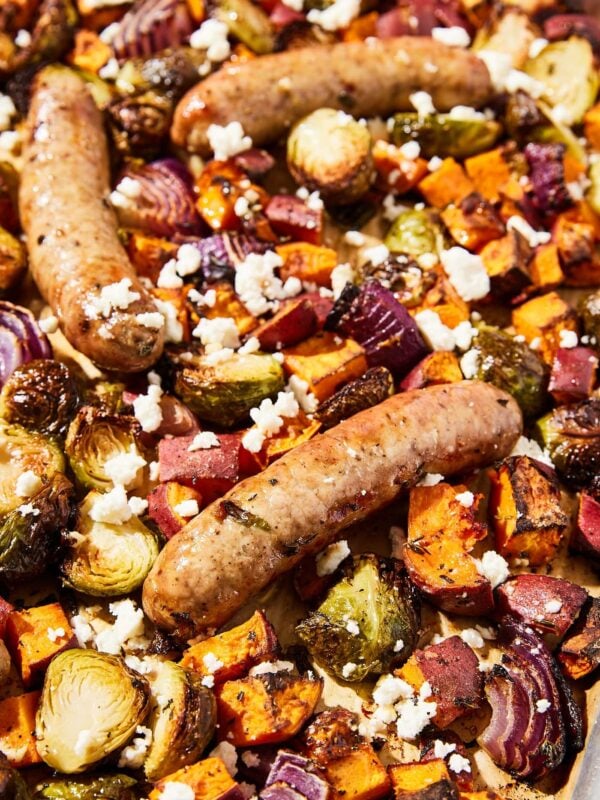 Large sheet pan with roasted vegetables and sausage links topped with vinaigrette and feta cheese.