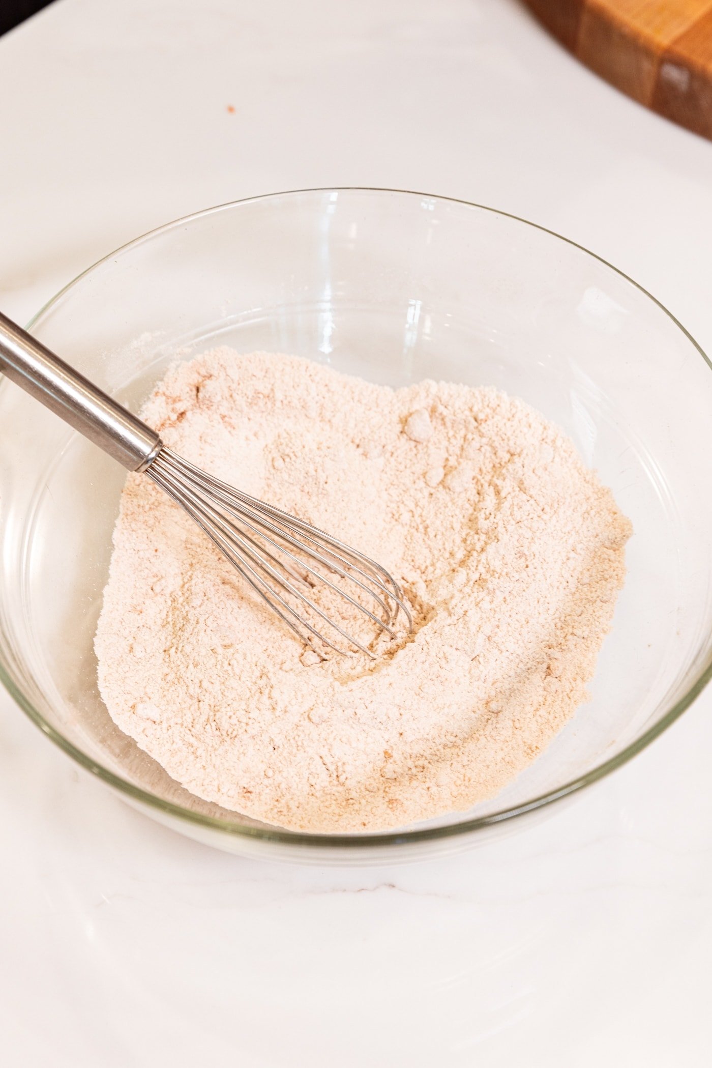Wire whisk resting against surface of glass bowl with whisked dry ingredients for carrot muffins.