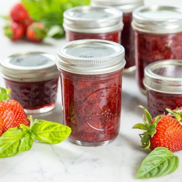 A jar of strawberry preserve sits on a table near basil and strawberries.