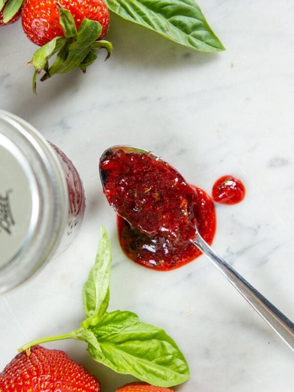 A spoon with strawberry preserve rests on a table next to a jar and strawberries.