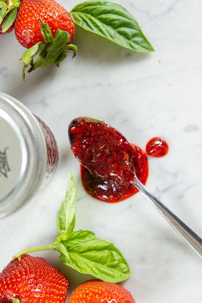 A spoon with strawberry preserve rests on a table next to a jar and strawberries.