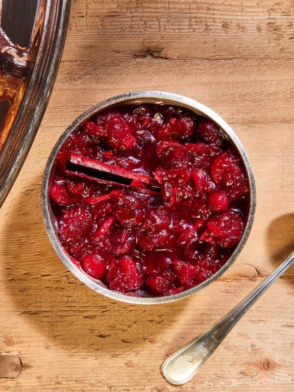 Silver bowl sitting on wooden surface filled with cranberry marmalade with cinnamon stick resting against side of bowl.