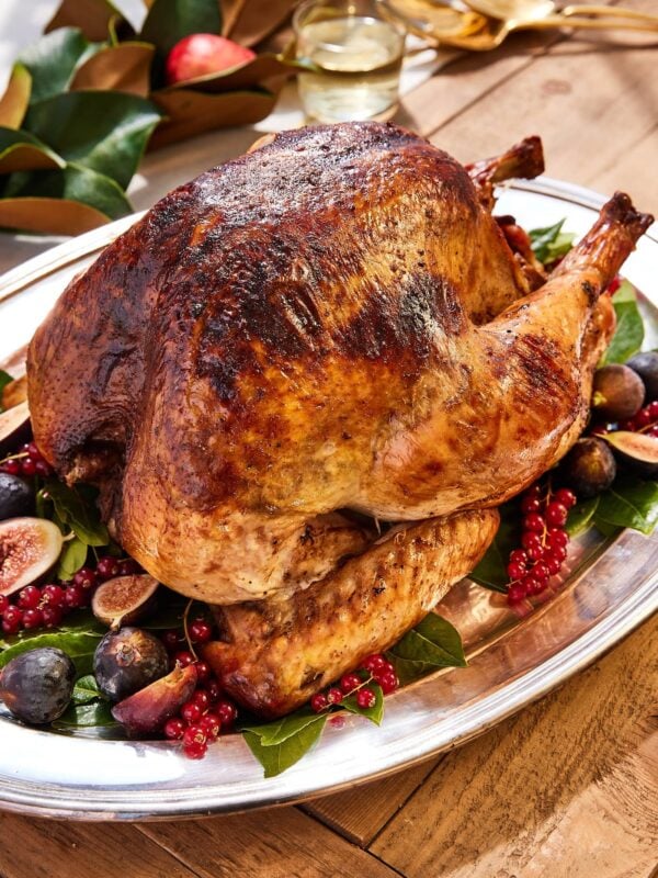 Large browned turkey sitting on silver platter with sliced figs and bay leaves underneath all on wooden table with wine glass in background.