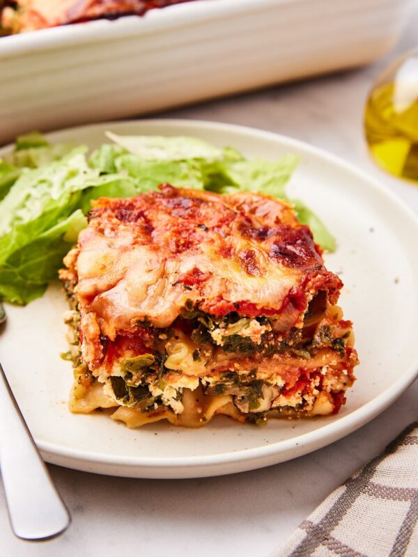 Cream color plate filled with square of vegetable lasagna and a side salad with rest of lasagna in the background.