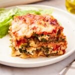 White plate with square of vegetable lasagna showing all interior layers including ricotta filling and vegetables with small salad behind.