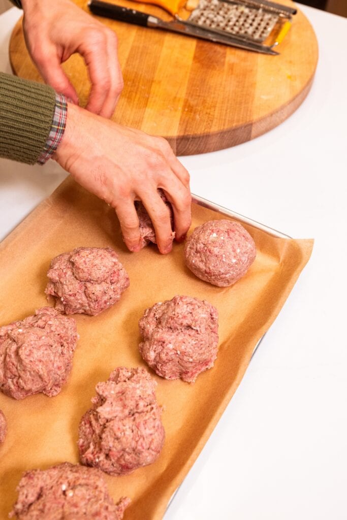 Parchment-lined baking sheet filled with rolled out meatballs all on white surface.