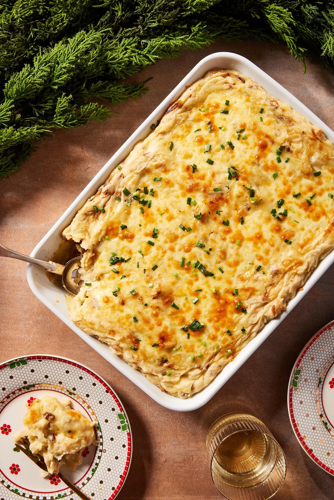 White baking dish filled with potato casserole with cheese melted on top sprinkled with parsley with serving on a Christmas plate with garland.