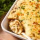 White baking dish filled with French onion potato casserole with cheese-covered topping with spoon scooping out a serving.