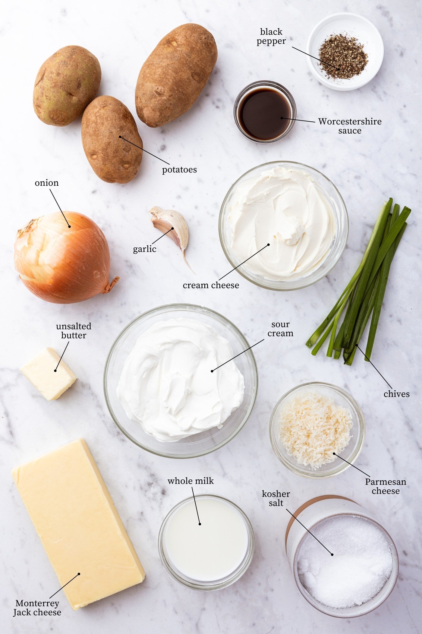 White marble surface filled with all ingredients needed to make French onion potato casserole including potatoes, sour cream, cream cheese, onions, cheese, and more.