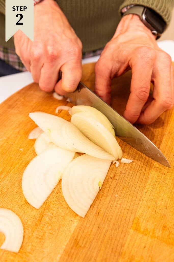 Two hands slicing onion into thin strips on wood cutting board with knife.