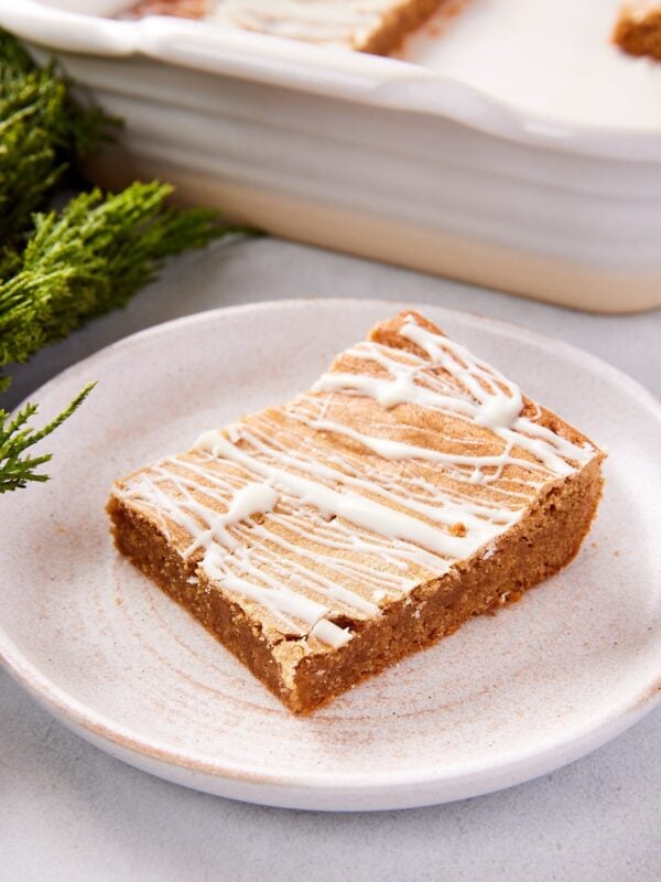 Stoneware plate with slice of gingerbread blondies with white chocolate drizzled on top with greenery in background.