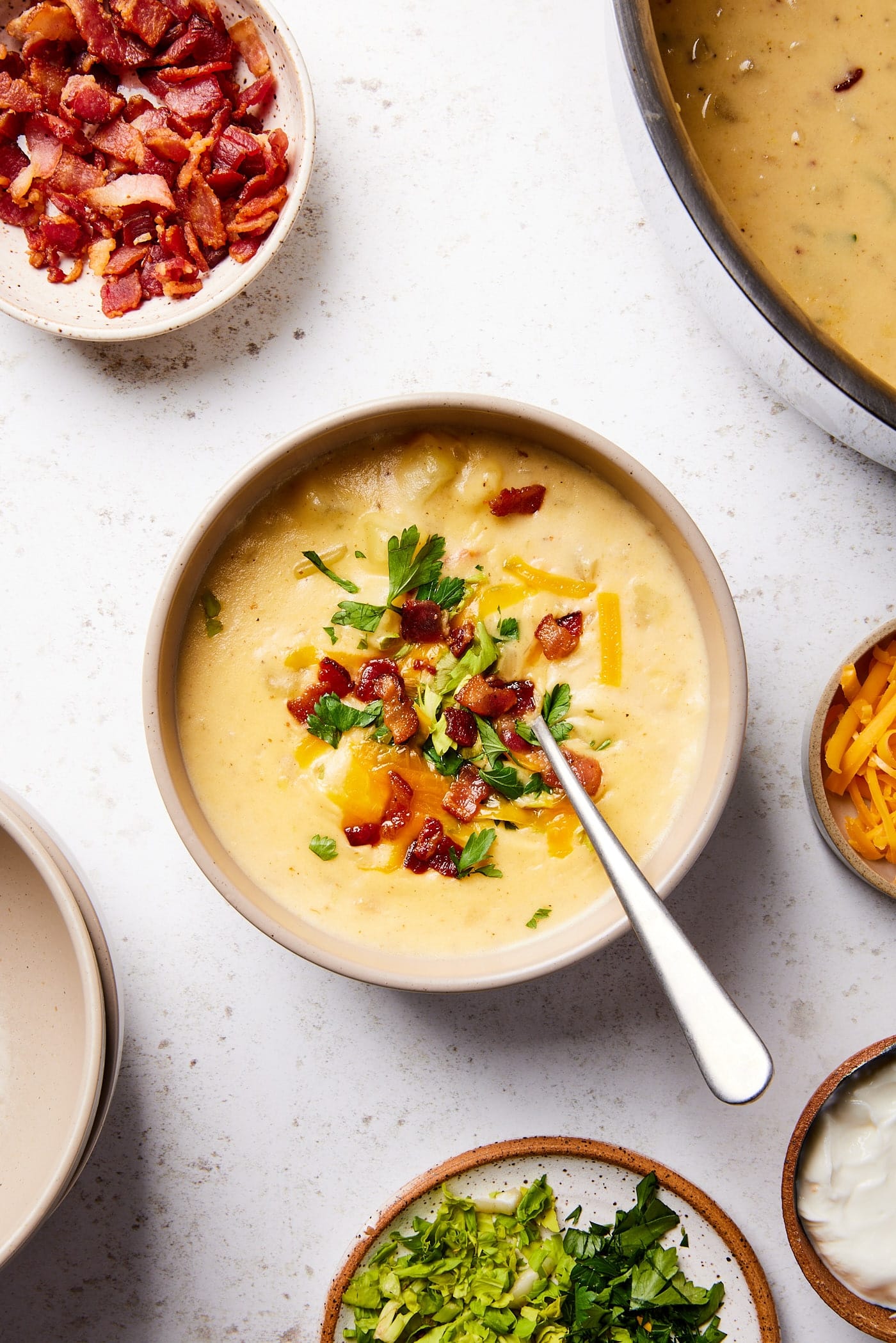 Bowl filled with serving of loaded baked potato soup with bacon and cheese sprinkled on top with rest of toppings on surface.