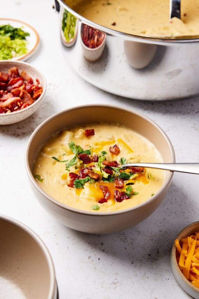 Brown bowl filled with loaded baked potato soup with bacon and cheese on top with spoon resting on side of bowl.