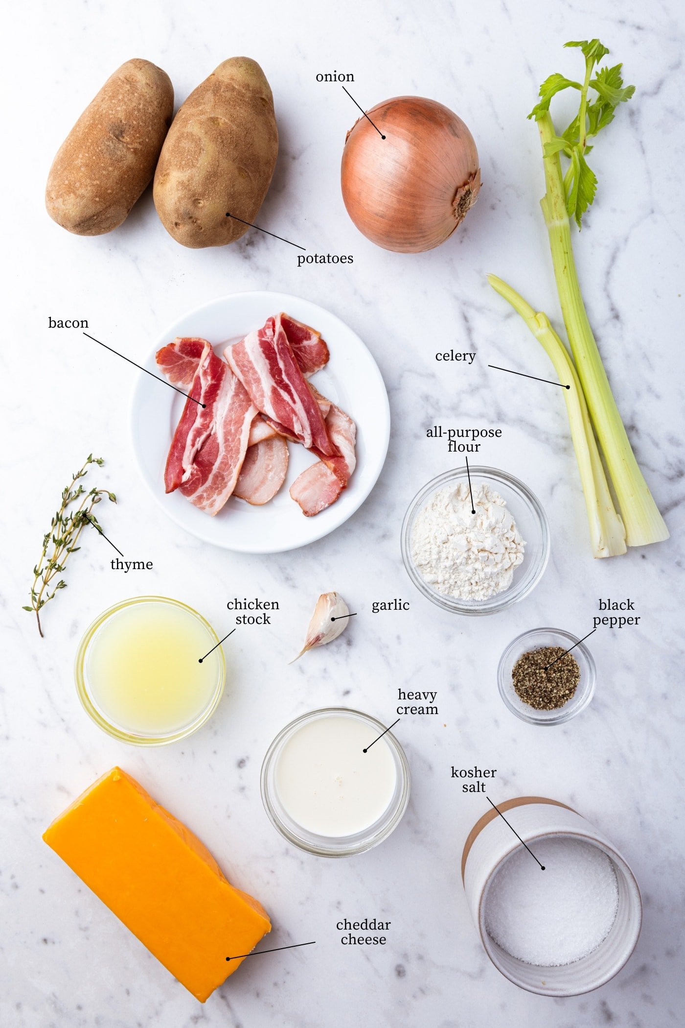White marble surface with all ingredients needed to make baked potato soup including potatoes, onion, celery, bacon, spices, and more.