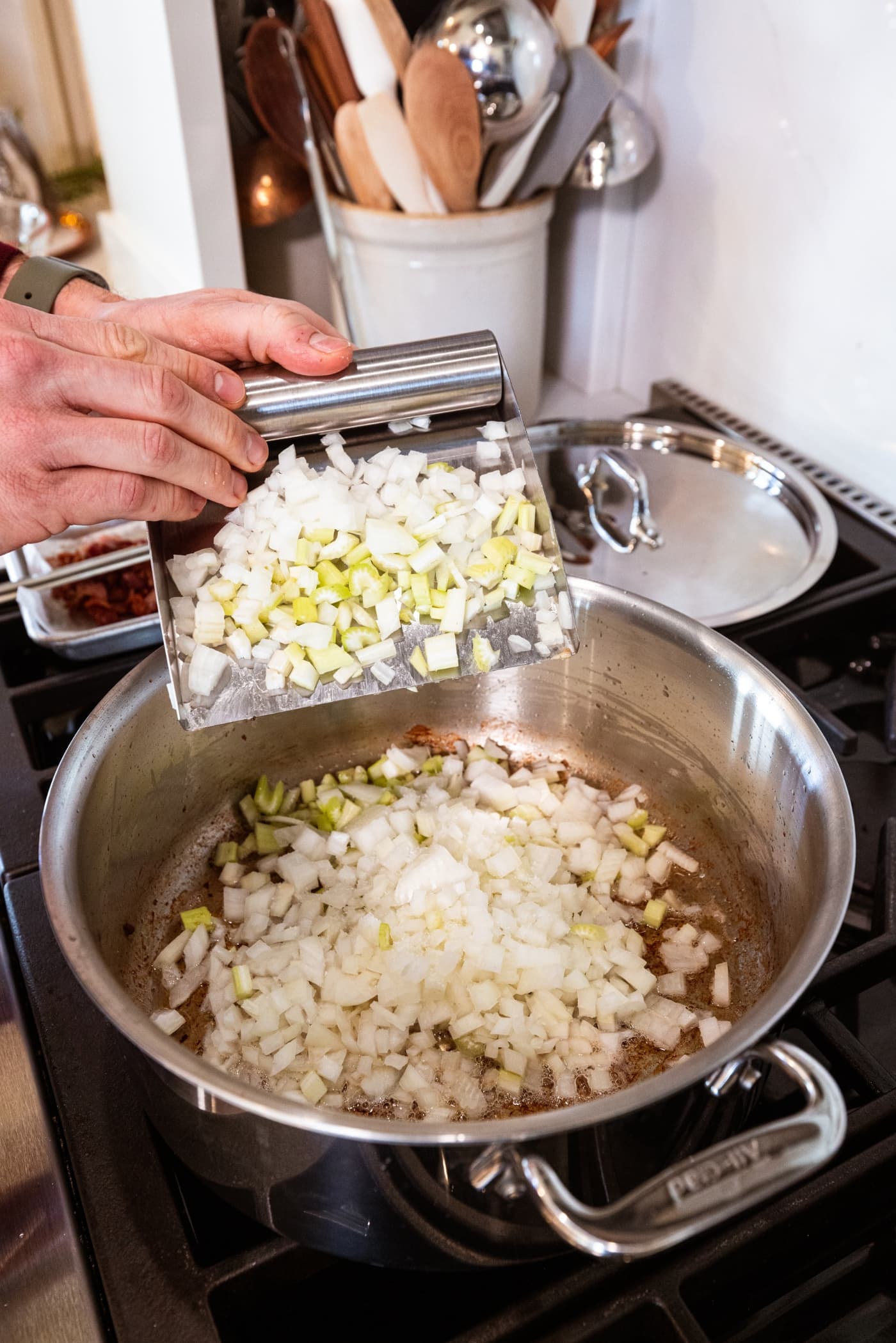 Hand using a scraper to throw diced onion and celery into a large stockpot on the stove.