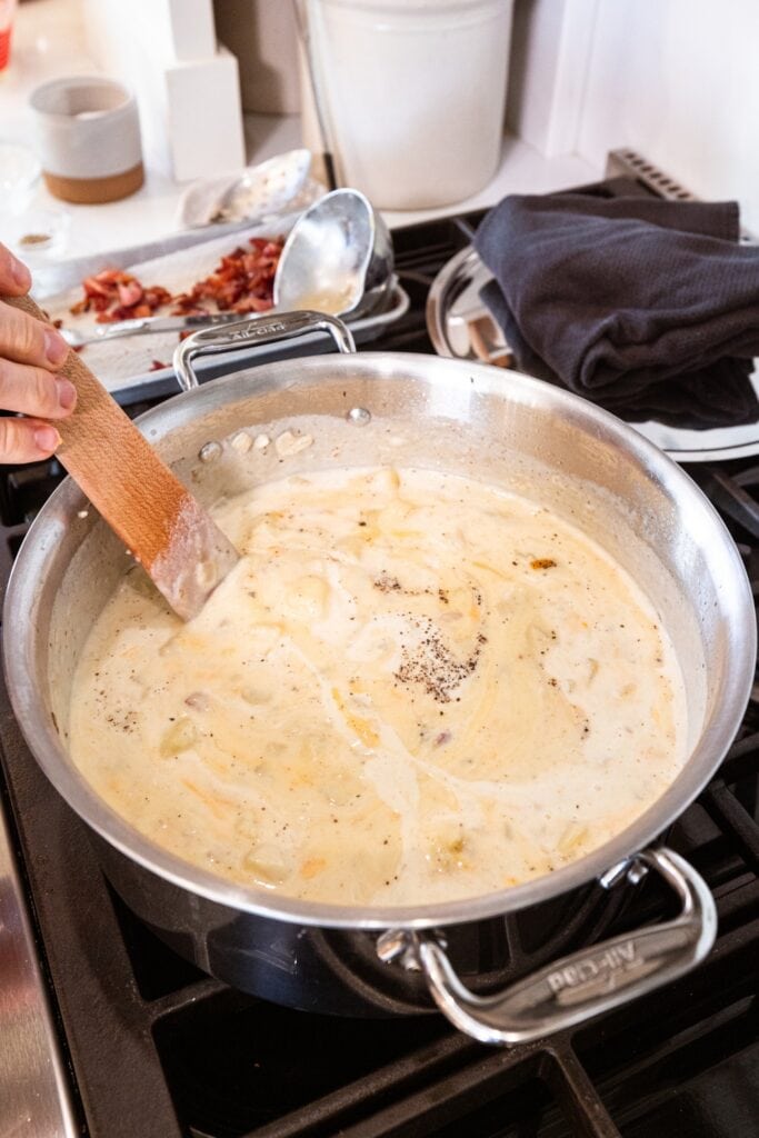 Wooden spatula stirring cream into loaded baked potato soup in large stockpot over stovetop.