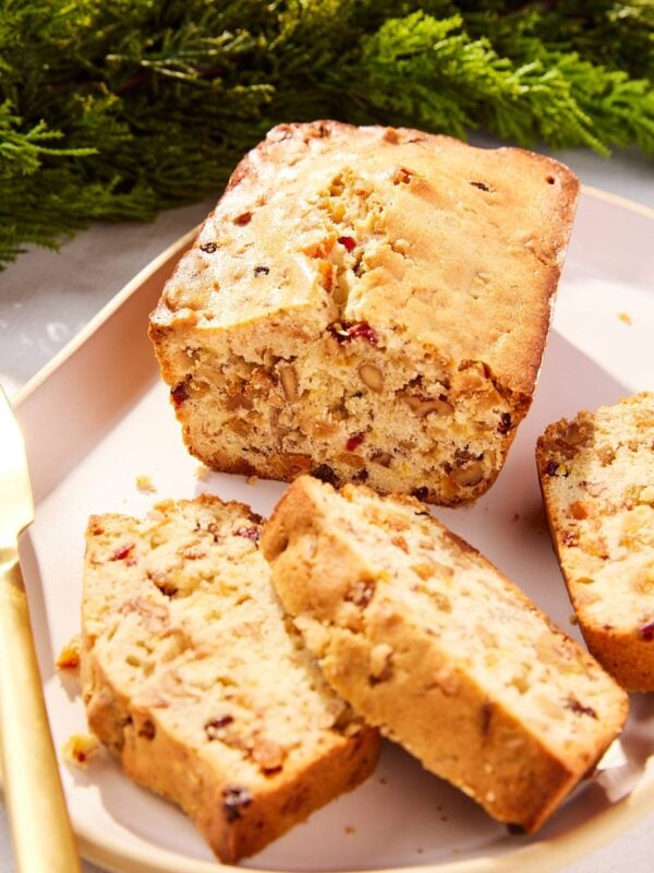 White fruitcake with slices sliced resting on a serving platter with gold knife and greenery in the background.
