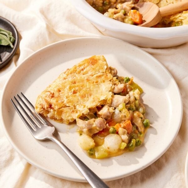 White plate resting on tablecloth with serving of chicken pot pie with fork resting on plate as well with more pot pie in dish behind.
