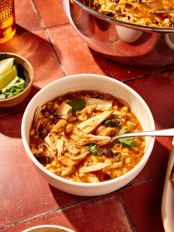 Bowl of chicken tortilla soup resting on red tile surface with more soup in the background and lots of toppings all around.