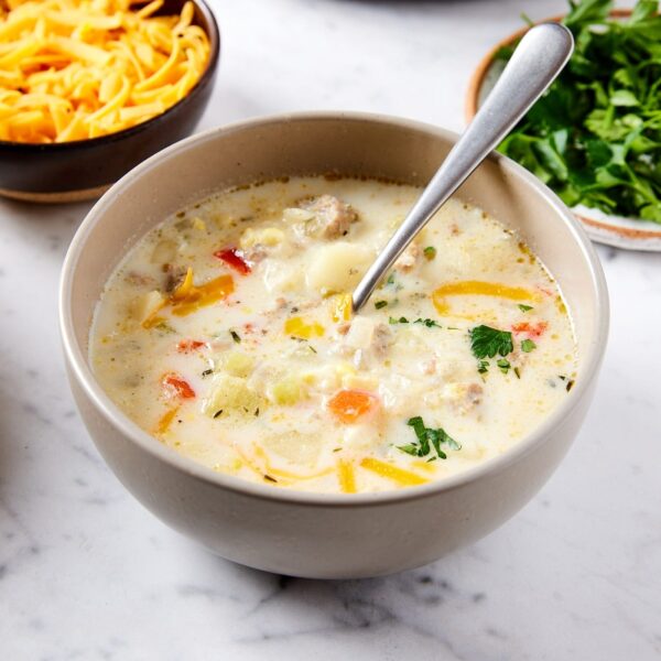 White marble surface with bowl of creamy corn chowder with shredded cheese and chopped parsley with spoon resting against side of bowl.
